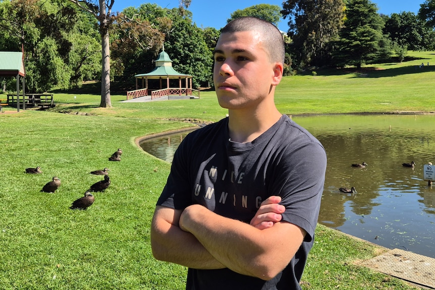 A teenage boy with close shaved hair in a grey tshirt stands with crossed arms in front of a lake in a green grassy park.