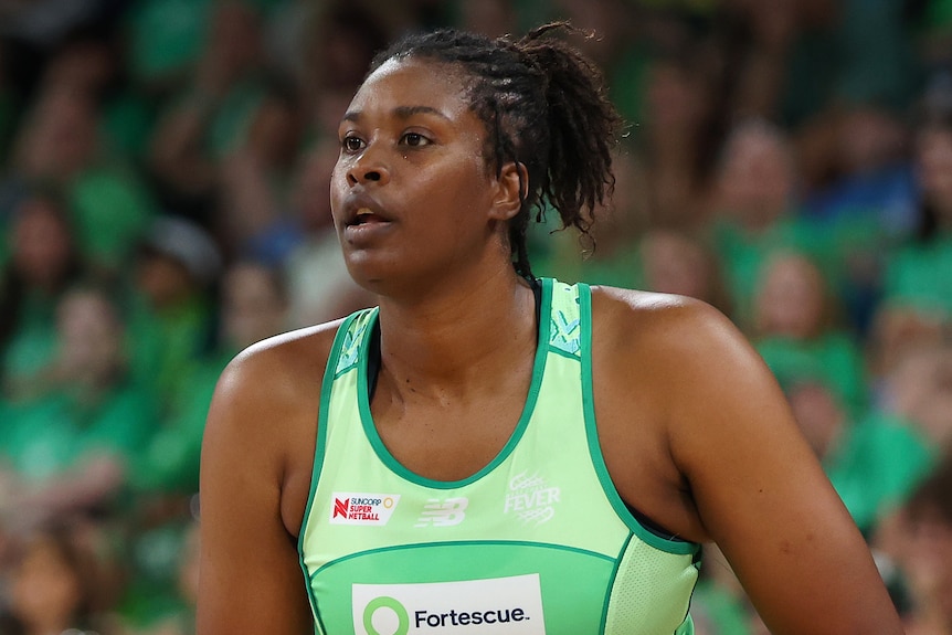 A player wears a green dress and holds a netball in front of her waist
