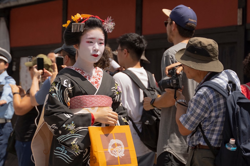 A woman made up in white face paint and red lipstick wearing a black kimono walks past a group of tourists.