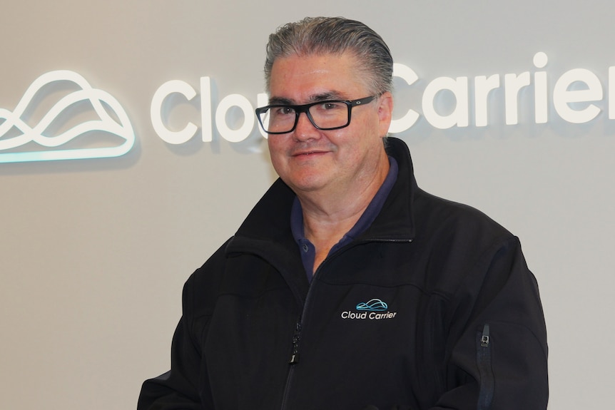 Greg, middle-aged Caucasian, smiles, stands in front of a wall with cloud carrier logo in white, wears black shirt and glasses.