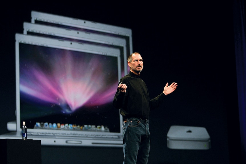 A man wearing a black turtleneck top stands on stage with his hands outstretched, a computer displayed on a screen behind him.