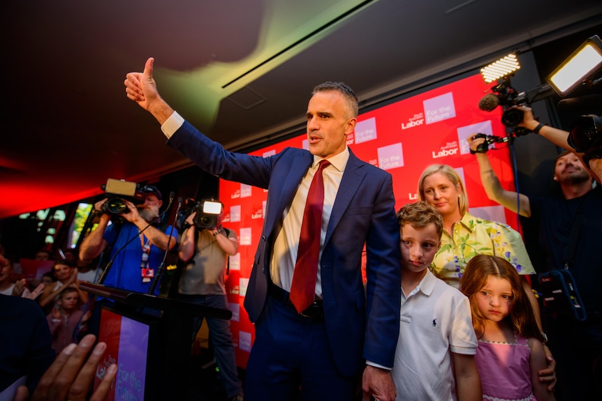 Peter Malinauskas gives a thumbs-up on stage next to his family in front of media cameras