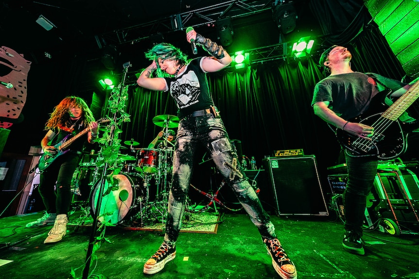 Des with green short hair stands on a stage in between two people playing guitars on a stage under green lights.