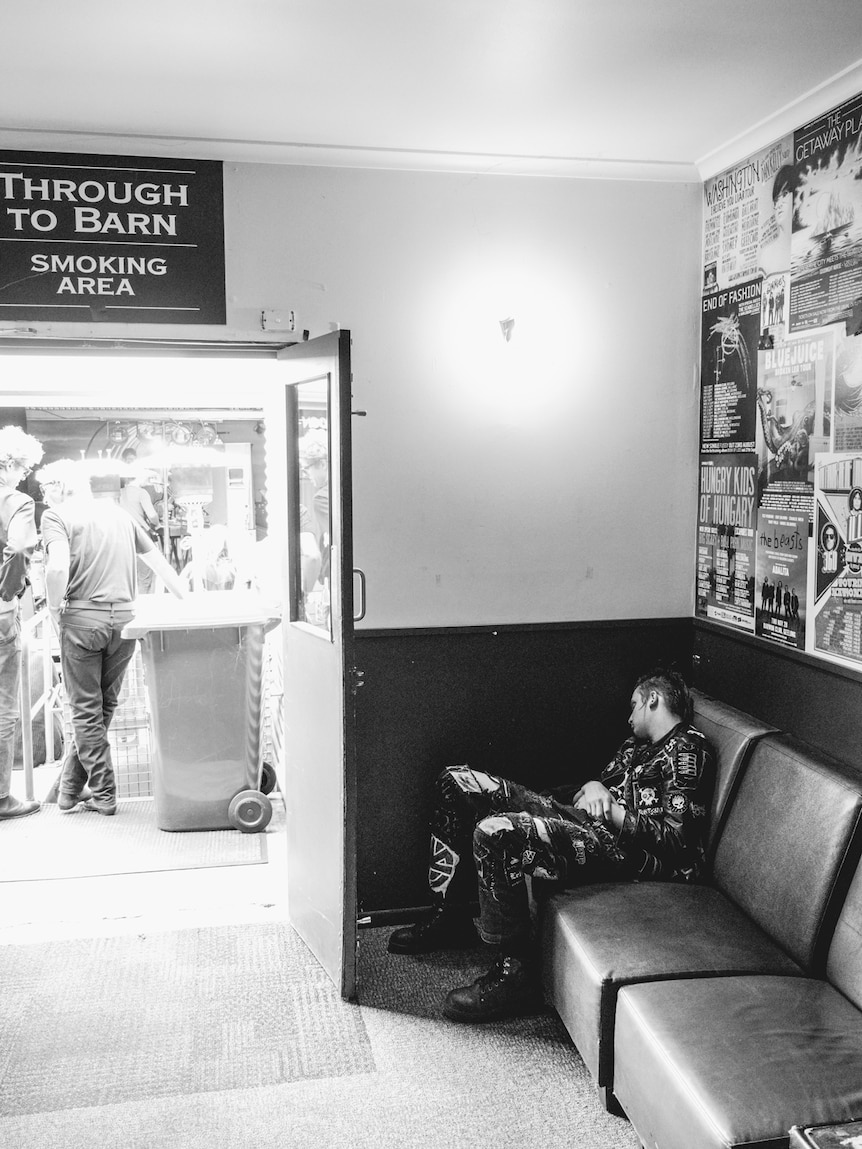 Black and white photo of a man in punk clothing napping in a corner of a live music venue next to an open door