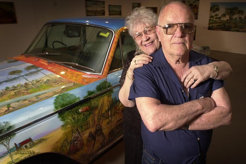 A woman and a man in front of a vintage car in a garage.