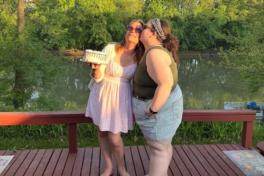 Rachel kisses Laura's cheek standing on a deck in front of a lake.