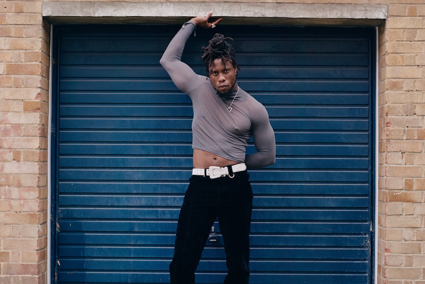 British-Nigerian man with dreads, wearing tight grey longsleeve, white belt and black jeans, poses in front of roller door