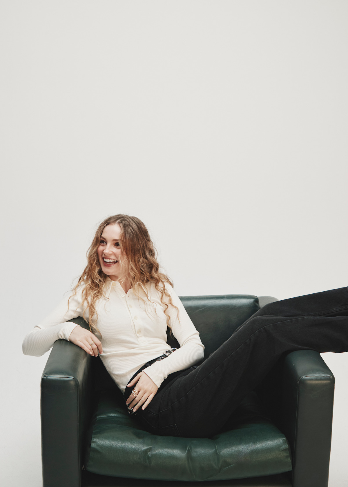 A smiling woman with long hair sits casually on a dark armchair, wearing a white long-sleeve top and black pants, with one leg draped over the armrest, in a minimalist studio with a light background.