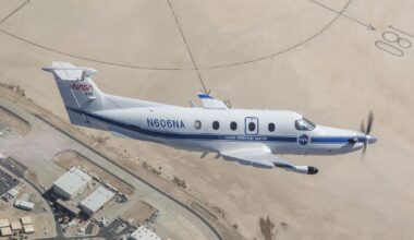 NASA’s freshly painted Pilatus PC-12 flies over NASA’s Armstrong Flight Research Center in Edwards, California. The parking lots and center buildings dot the landscape below. The compass rose in the upper part of the photo shaped like the sun hovers over the aircraft, emphasizing the gleaming white plane with a blue stripe and blue N606NA number across the fuselage and NASA red worm logo on the tail. A sensor, with a black-tip juts out from below the wing.