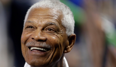 A smiling elderly man with short white hair and a mustache wears a white shirt, appearing joyful and looking slightly upward.