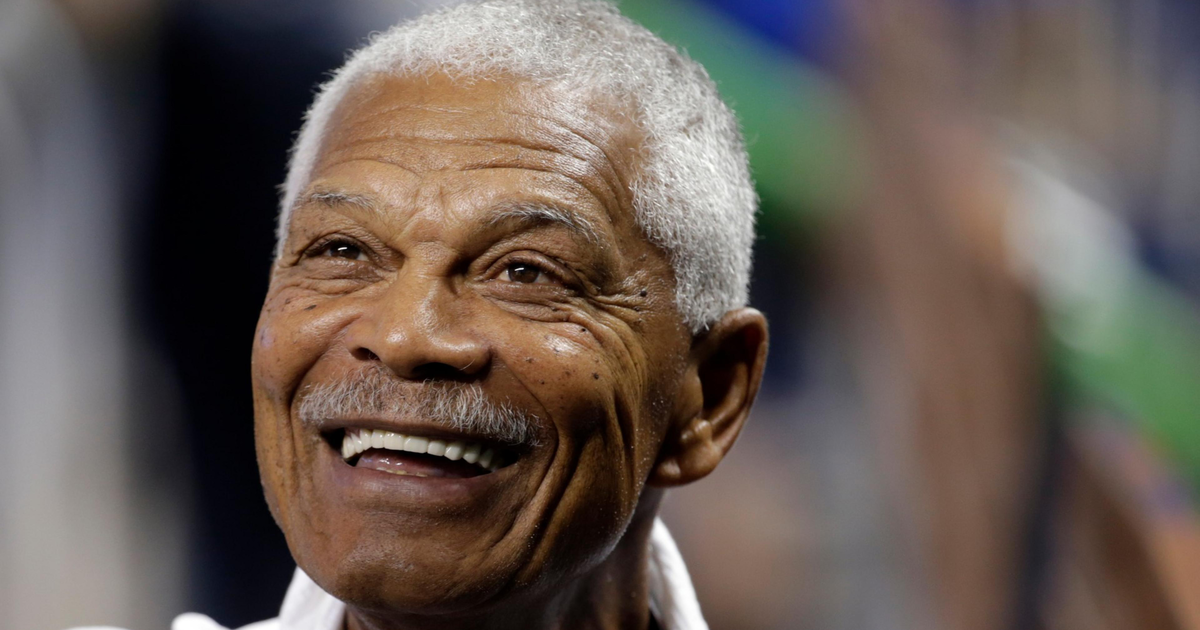 A smiling elderly man with short white hair and a mustache wears a white shirt, appearing joyful and looking slightly upward.