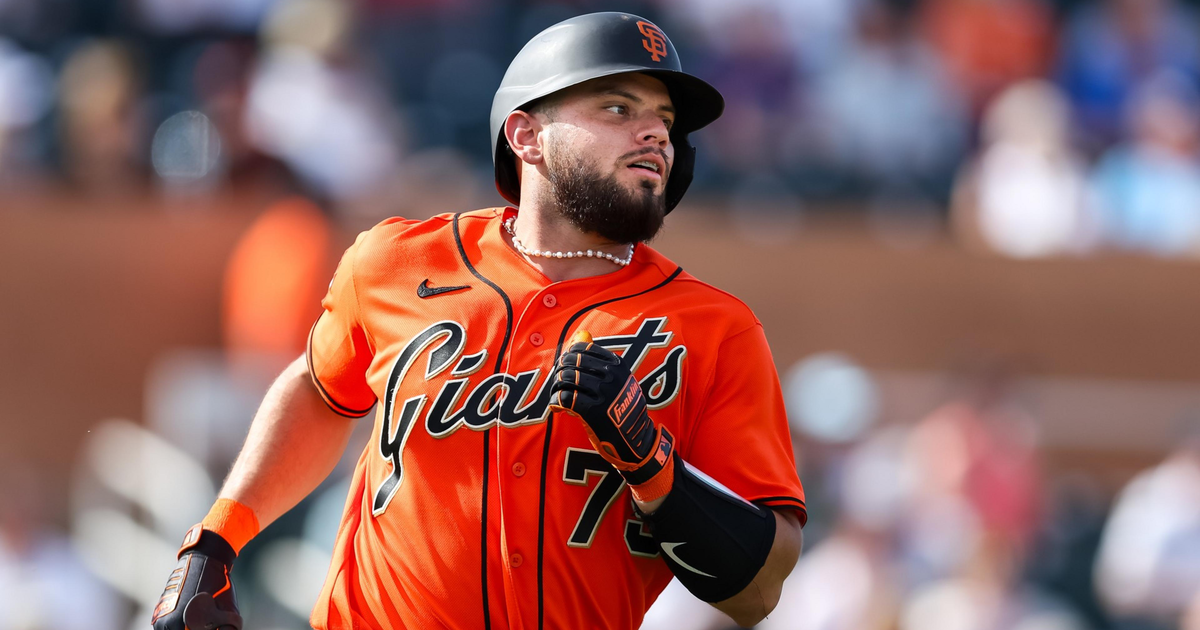 A baseball player in a bright orange San Francisco Giants uniform runs, wearing a black helmet, batting gloves, and a pearl necklace.
