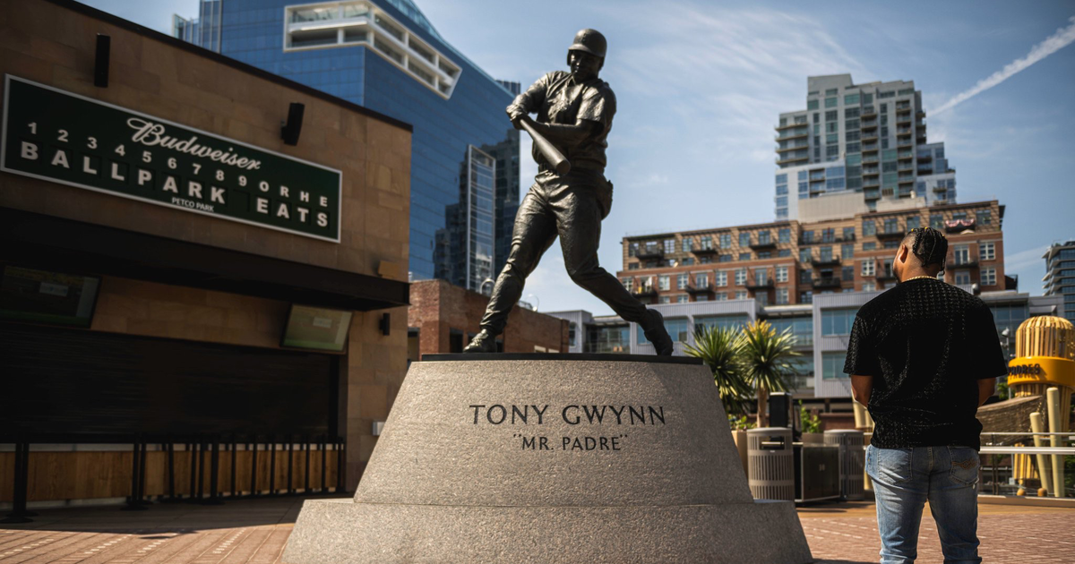 A bronze statue of baseball player Tony Gwynn in batting stance stands on a pedestal labeled "Mr. Padre" with a man looking at it nearby.