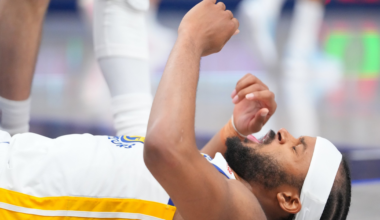 A basketball player wearing a white and yellow jersey lies on the floor with eyes closed and hands raised, possibly in pain or exhaustion.