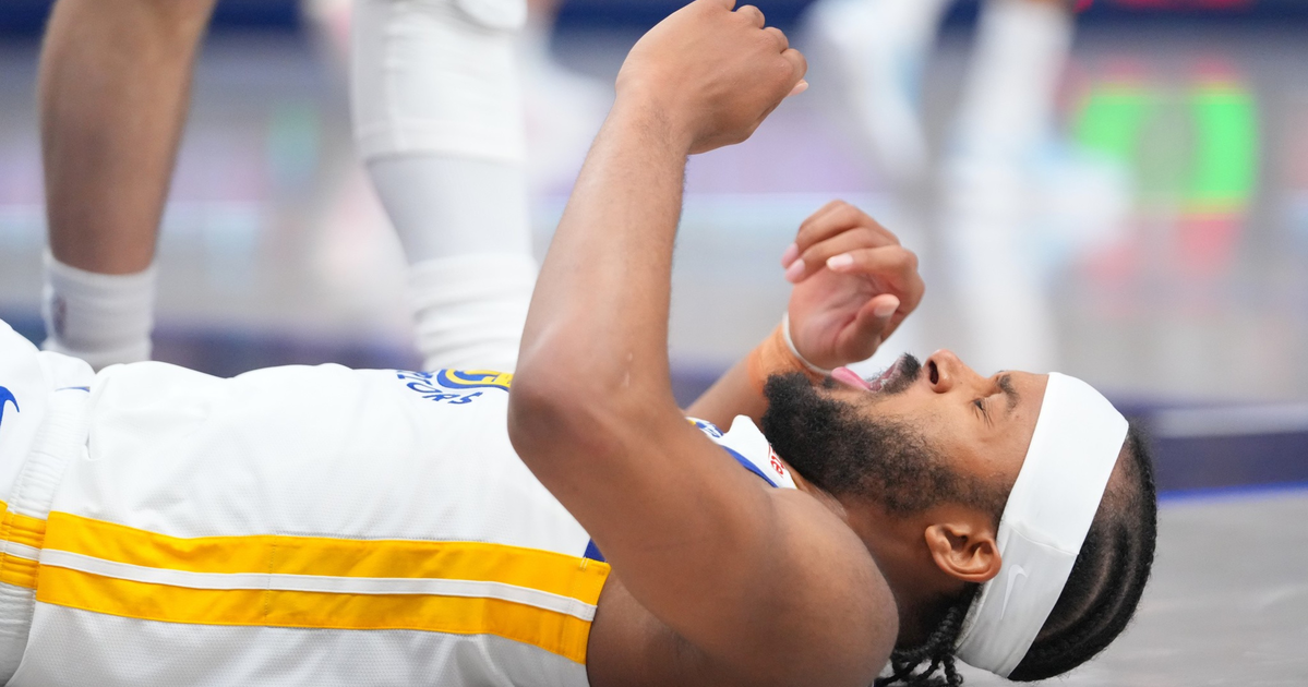 A basketball player wearing a white and yellow jersey lies on the floor with eyes closed and hands raised, possibly in pain or exhaustion.