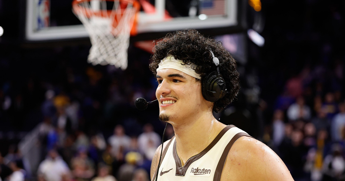 A basketball player wearing a white "The Town" jersey and headset smiles on the court with a basketball hoop and crowd blurred in the background.