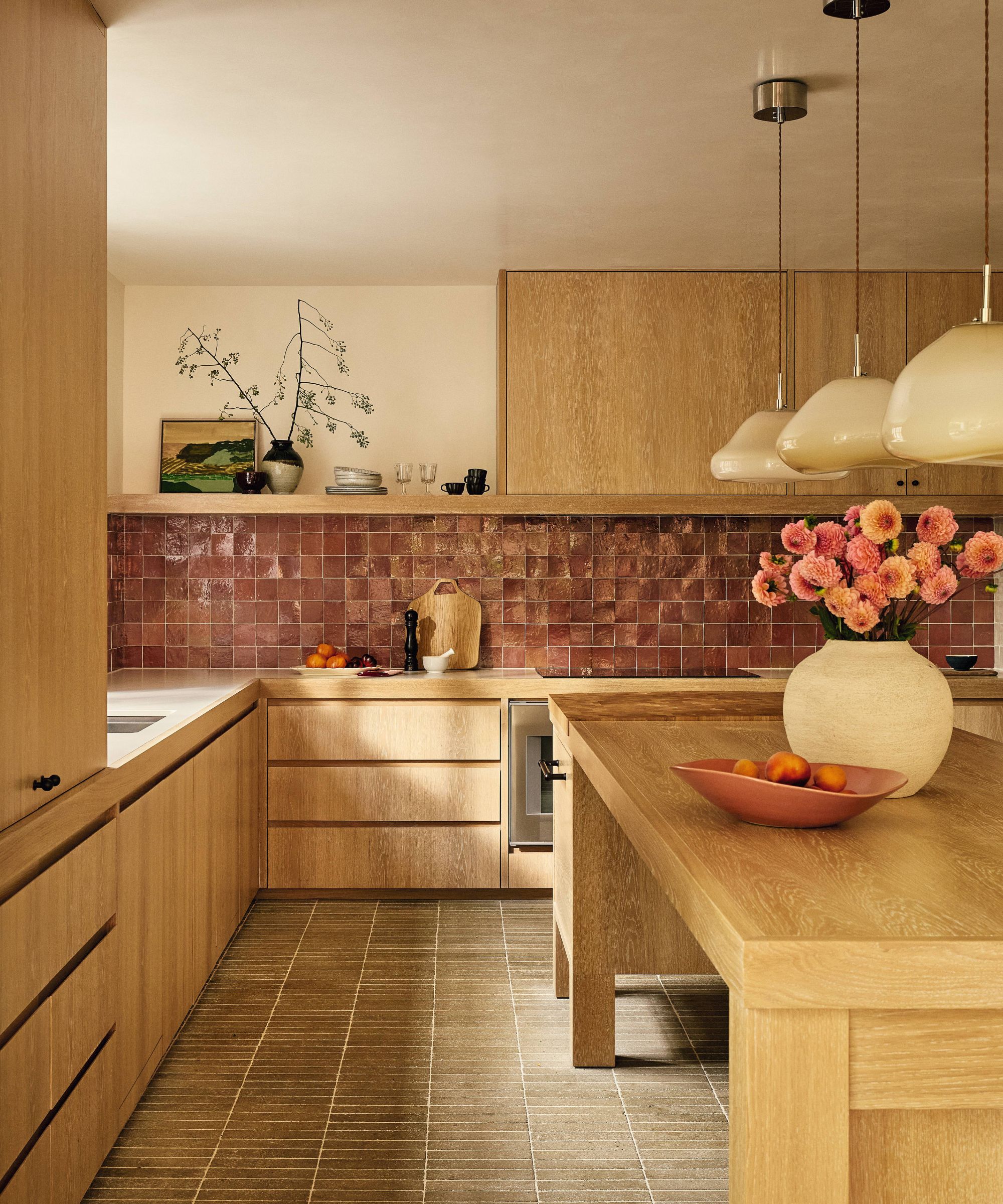 a warm modern kitchen with oak cabinetry, green tiled floor, mauve zellige wall tiles and a large table island with pendants hung above