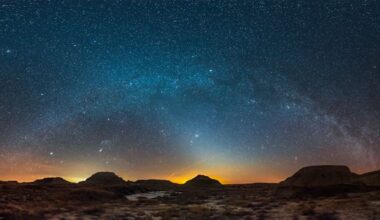 The soft glow of zodiacal light shines beneath the Milky way in a starry sky above an arid, rocky desert landscape.