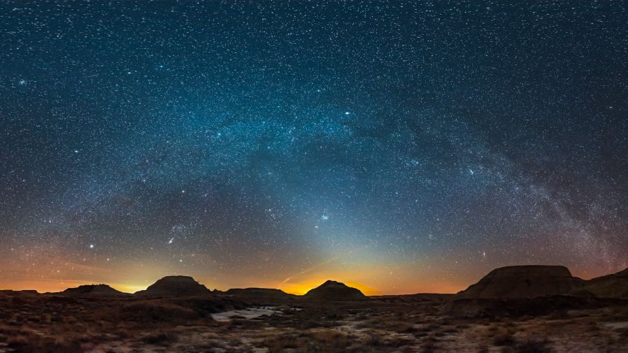 The soft glow of zodiacal light shines beneath the Milky way in a starry sky above an arid, rocky desert landscape.