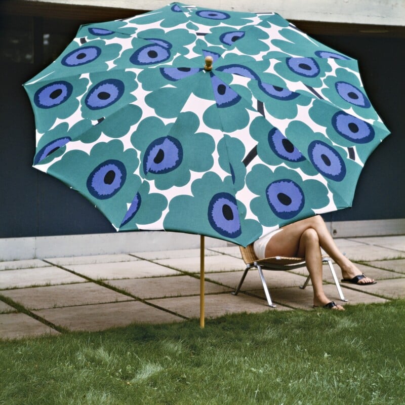 A person sits on a lounge chair with legs crossed, mostly hidden behind a large, colorful umbrella with a bold blue and green floral pattern, set on a patio next to grass.