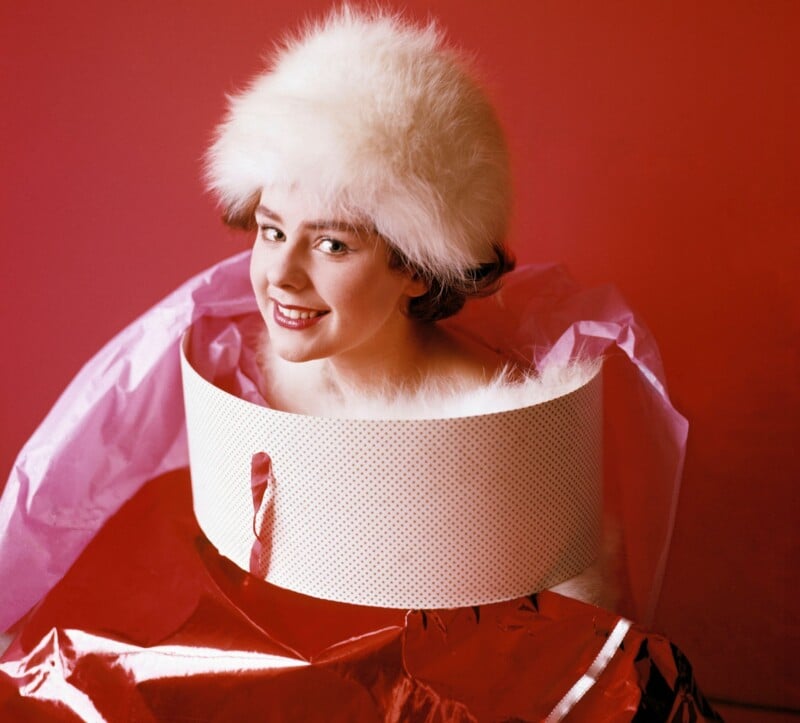 A smiling woman wearing a white fur hat and wrapped in red and pink shiny material poses against a red background. She also has a large, white, circular collar around her shoulders.