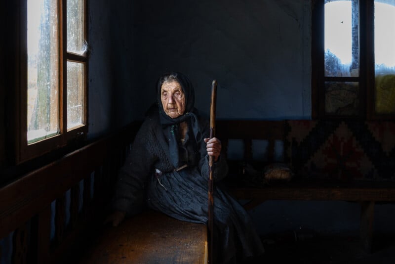 An elderly woman wearing a dark headscarf and coat sits on a wooden bench, holding a walking stick. She looks thoughtfully out the window as soft light illuminates her face. The room is dim and rustic.