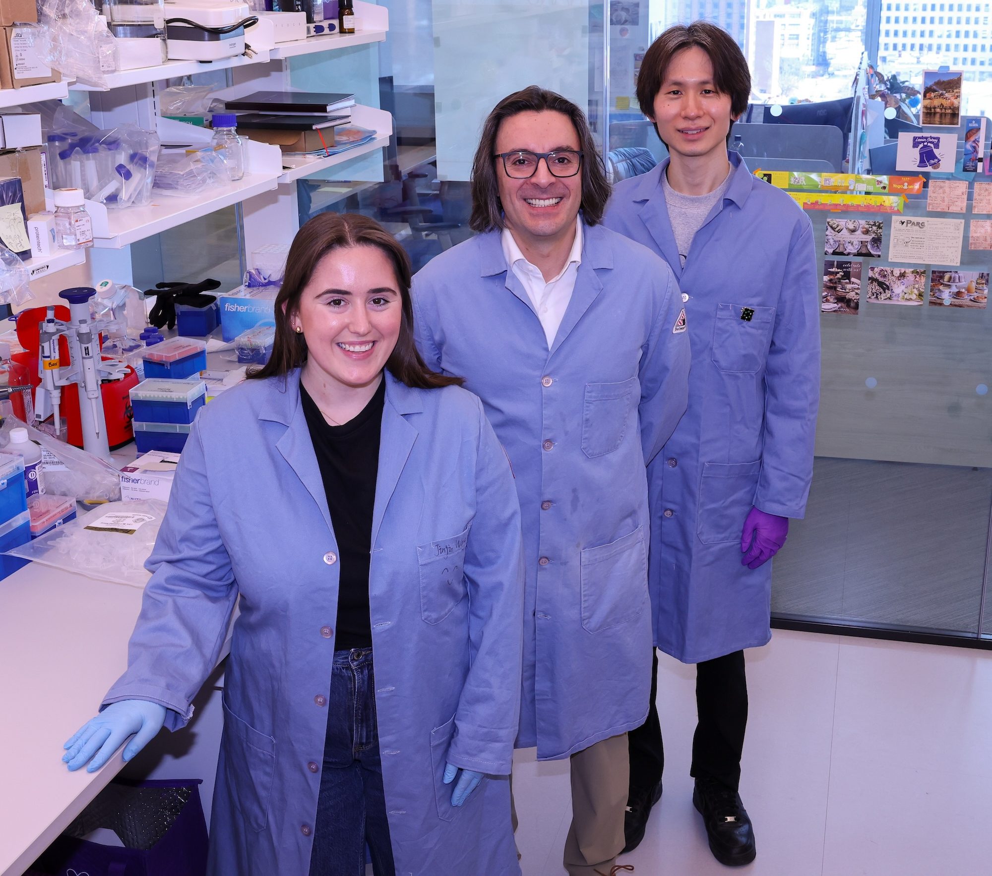 Three researchers in lab coats stand in a row next to a lab bench. 