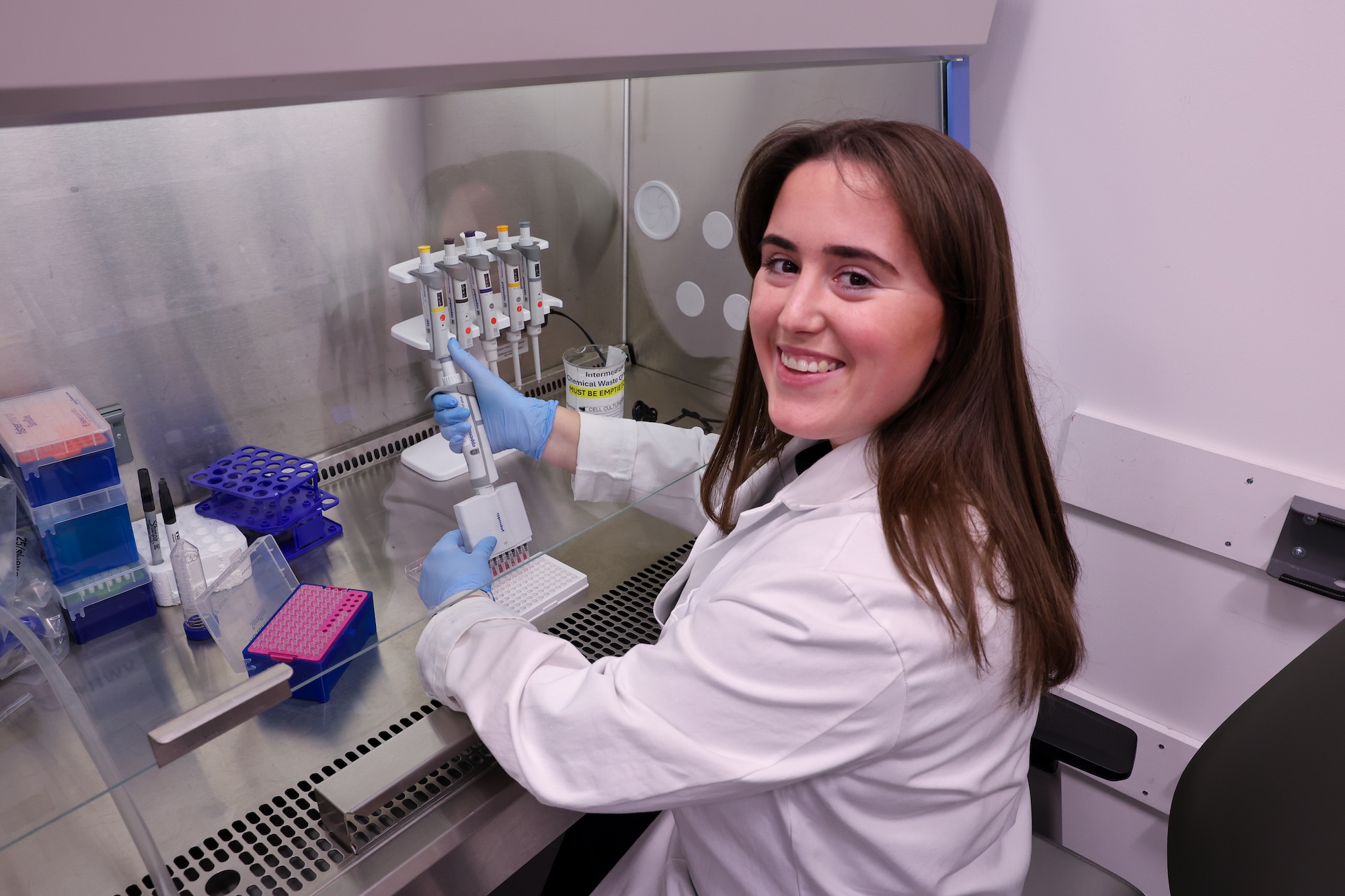 A woman in a lab coat looks toward the camera while holding a micropipette. 