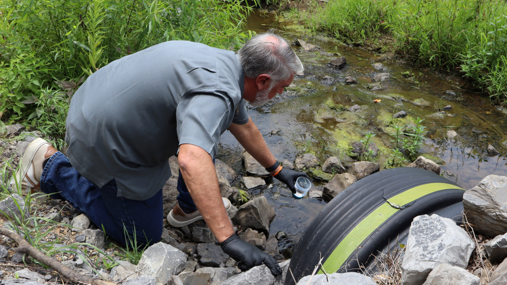 A man crouches in a stream and takes a water quality sample in a cup.