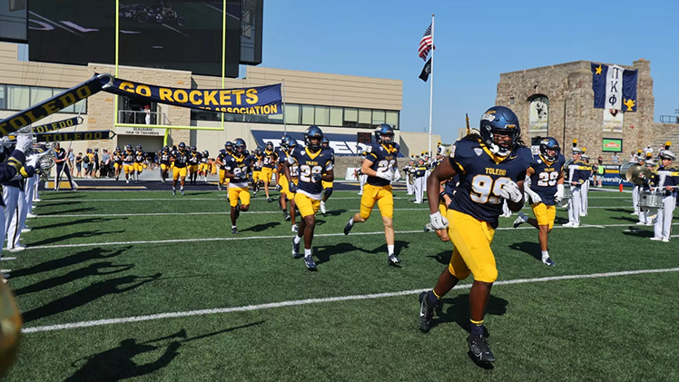 Toledo football players run onto the Glass Bowl field as the band plays next to them.