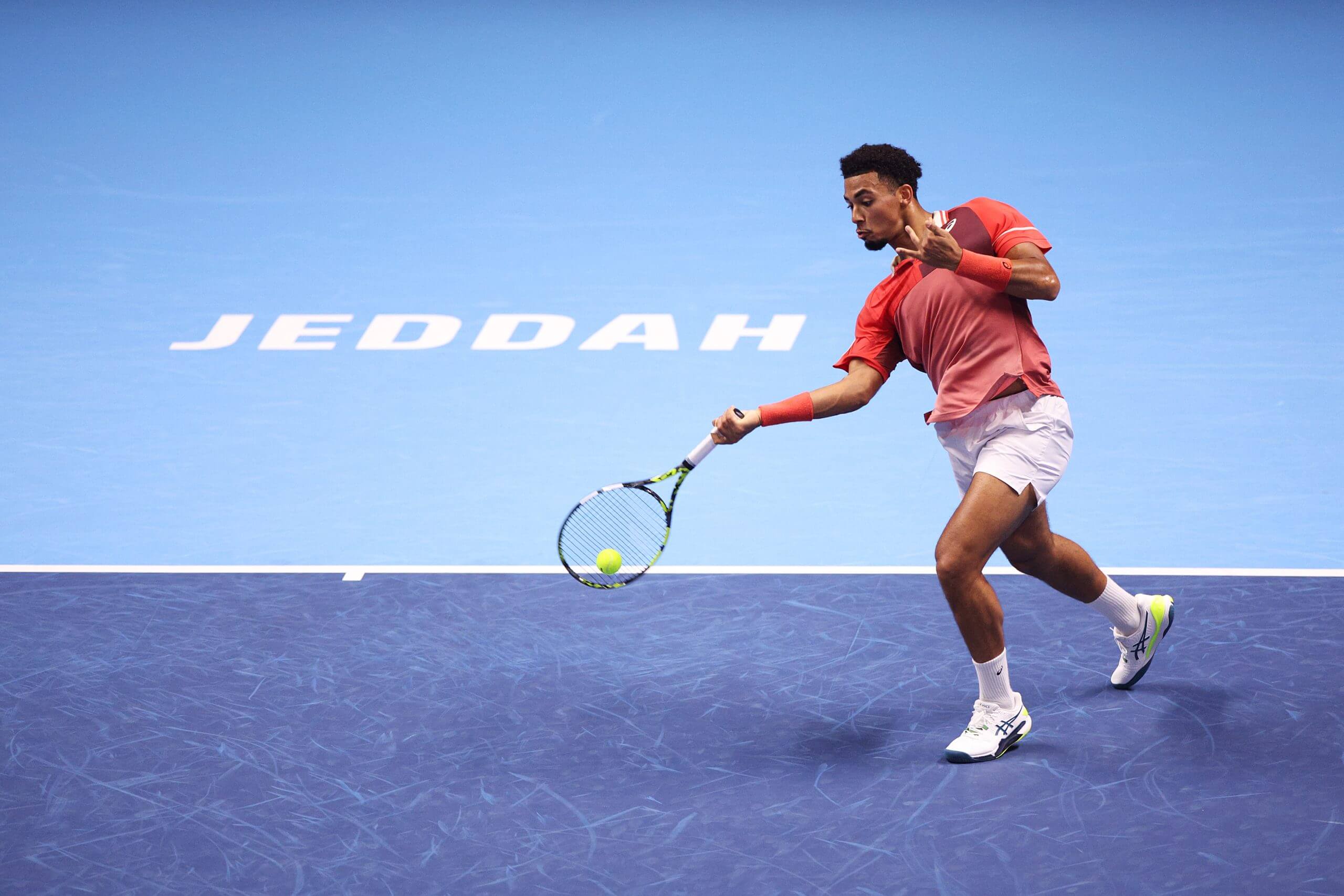 Arthur Fils hits a low forehand on a blue hard court, with “Jeddah” written in all-white capital letters on the light blue behind the baseline.