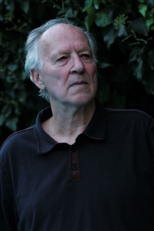 An older man with gray hair, wearing a dark collared shirt, stands outdoors in front of leafy green foliage, looking thoughtfully into the distance.