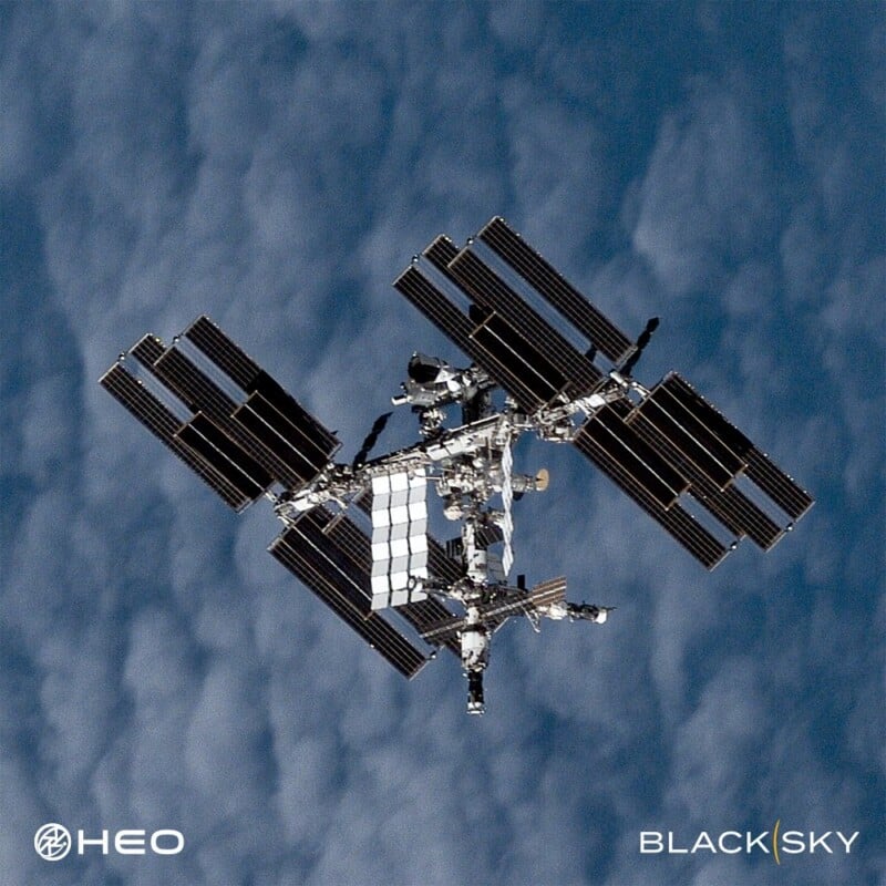 A detailed view of the International Space Station orbiting above Earth, with its solar panels extended; clouds and the blue atmosphere are visible in the background. Logos for HEO and BlackSky are in the corners.