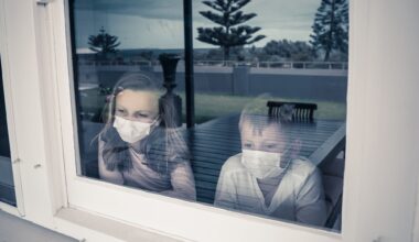 COVID-19 Lockdowns. Sad children with face mask looking through the window during quarantine at home. Kids feeling sad as school is closed. Coronavirus outbreak isolation and kids mental health.