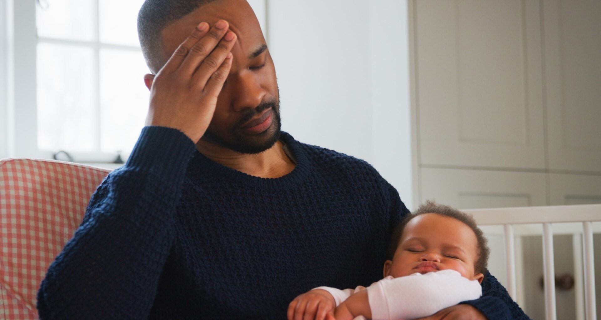 Stressed New Father Sitting In Chair Holding Sleeping Baby Girl In Nursery At Home