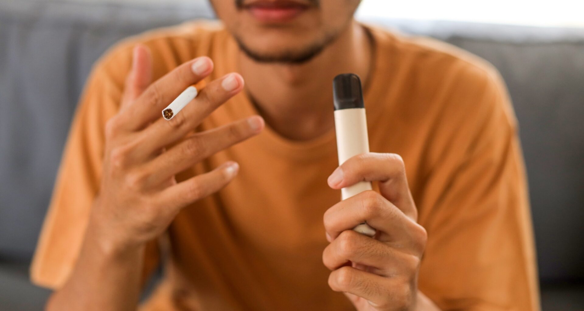 Young Asian man holding a cigarette and e-cigarette vaping device while sitting in couch