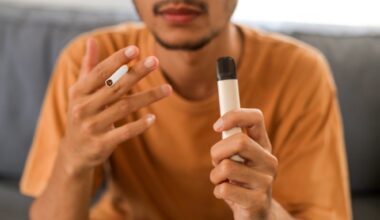 Young Asian man holding a cigarette and e-cigarette vaping device while sitting in couch
