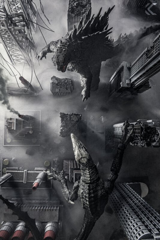 An aerial view of two giant monsters, resembling Godzilla and a reptilian creature, battling in a city surrounded by smoke, damaged buildings, and industrial structures.