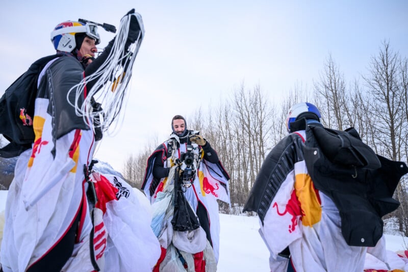 Three people wearing wingsuits with Red Bull logos stand in the snow, holding parachutes and gear, with bare trees and a pale sky in the background.