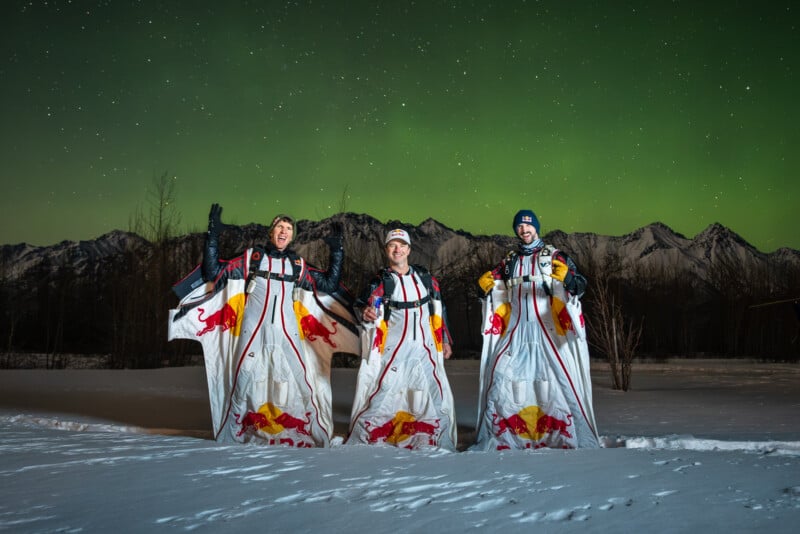Three people in Red Bull branded wingsuits pose on snowy ground at night, with mountains in the background and a green aurora borealis lighting up the sky above them.