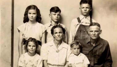A black-and-white portrait of a family from the early 20th century, showing two adults and four children. The adults are seated in front, with the children standing and sitting around them, all facing the camera with neutral expressions.