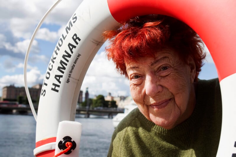 An older woman with red hair smiles while leaning through a life preserver labeled "Stockholms Hamnar" by the water, with a cityscape and partly cloudy sky in the background.