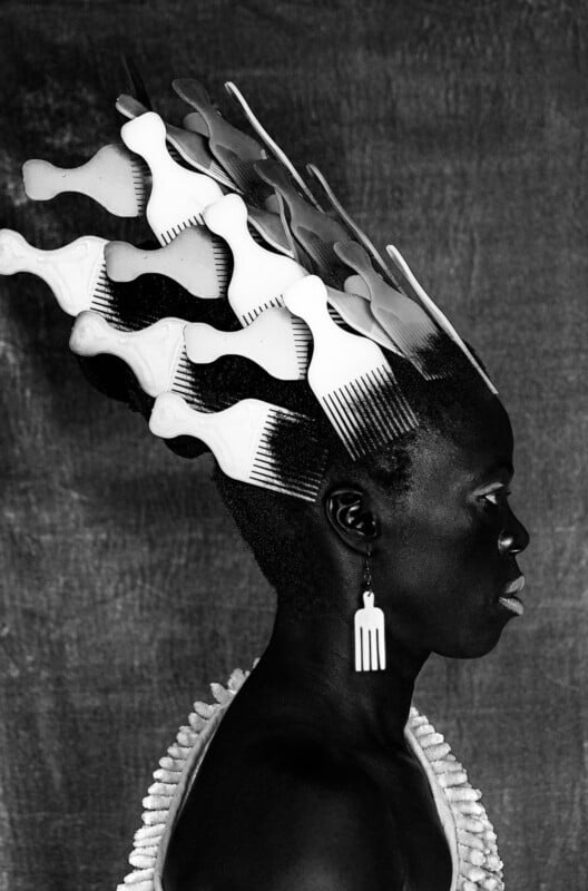 A Black person in profile with hair styled upward, adorned with multiple white Afro picks. They wear a comb-shaped earring and a textured top, set against a plain, textured background.