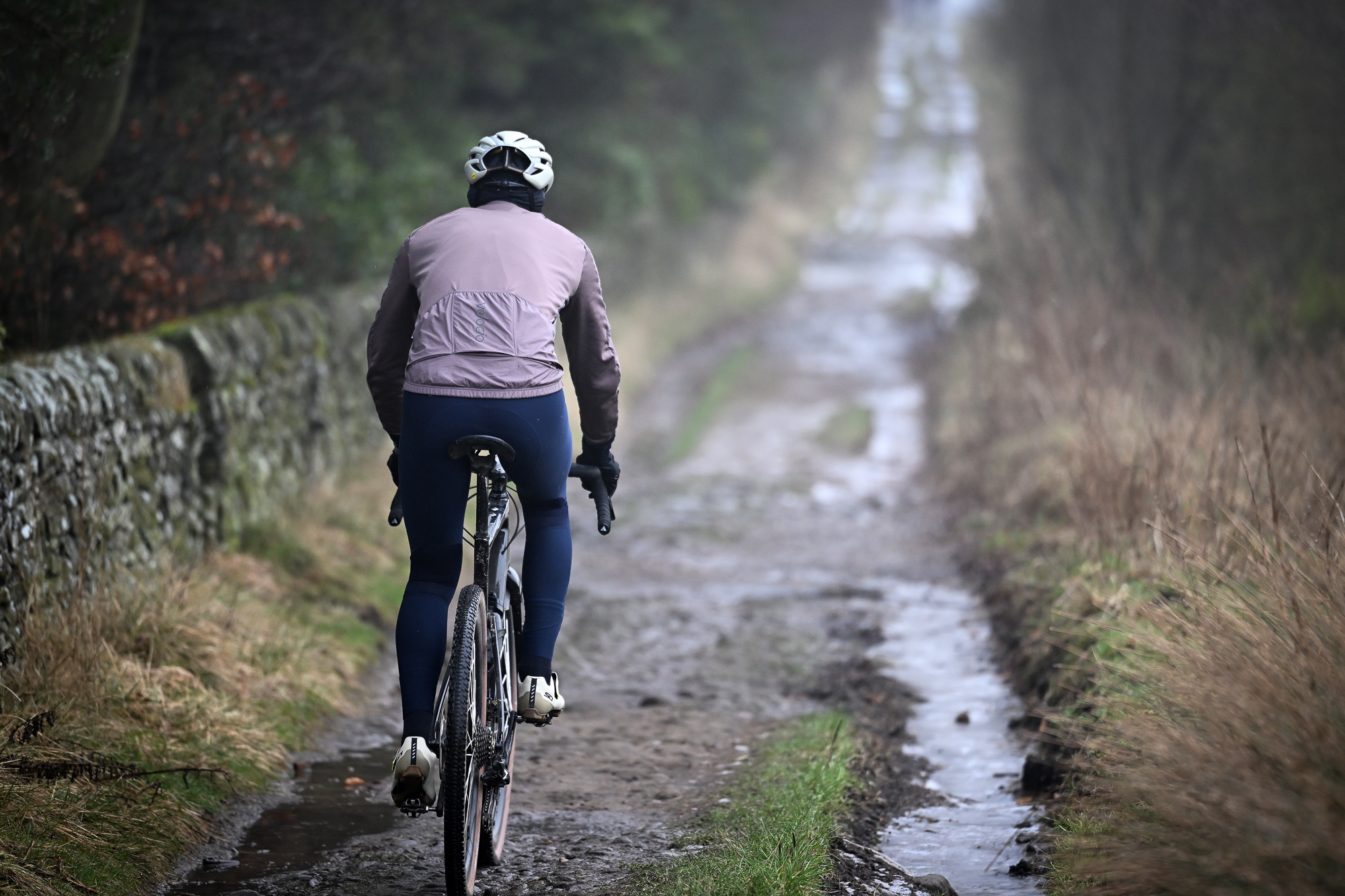 Man wearing a purple jacket, navy tights and a pale helmet riding a black gravel bike away from the camera on a muddy track