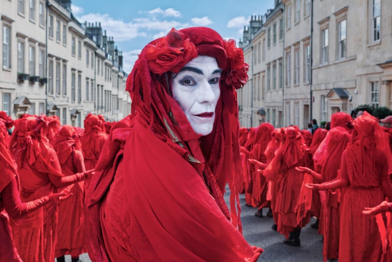 A person in white face paint and a red costume with a headpiece of red flowers stands in the foreground, surrounded by a group of people dressed entirely in red on a city street.