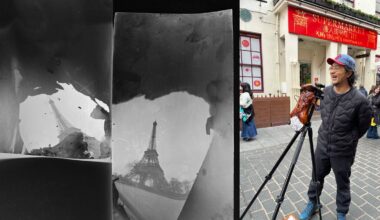 A man wearing a cap stands on a city street beside a large camera on a tripod. Next to him are two black-and-white photographic negatives, one showing the Eiffel Tower. A supermarket is visible in the background.
