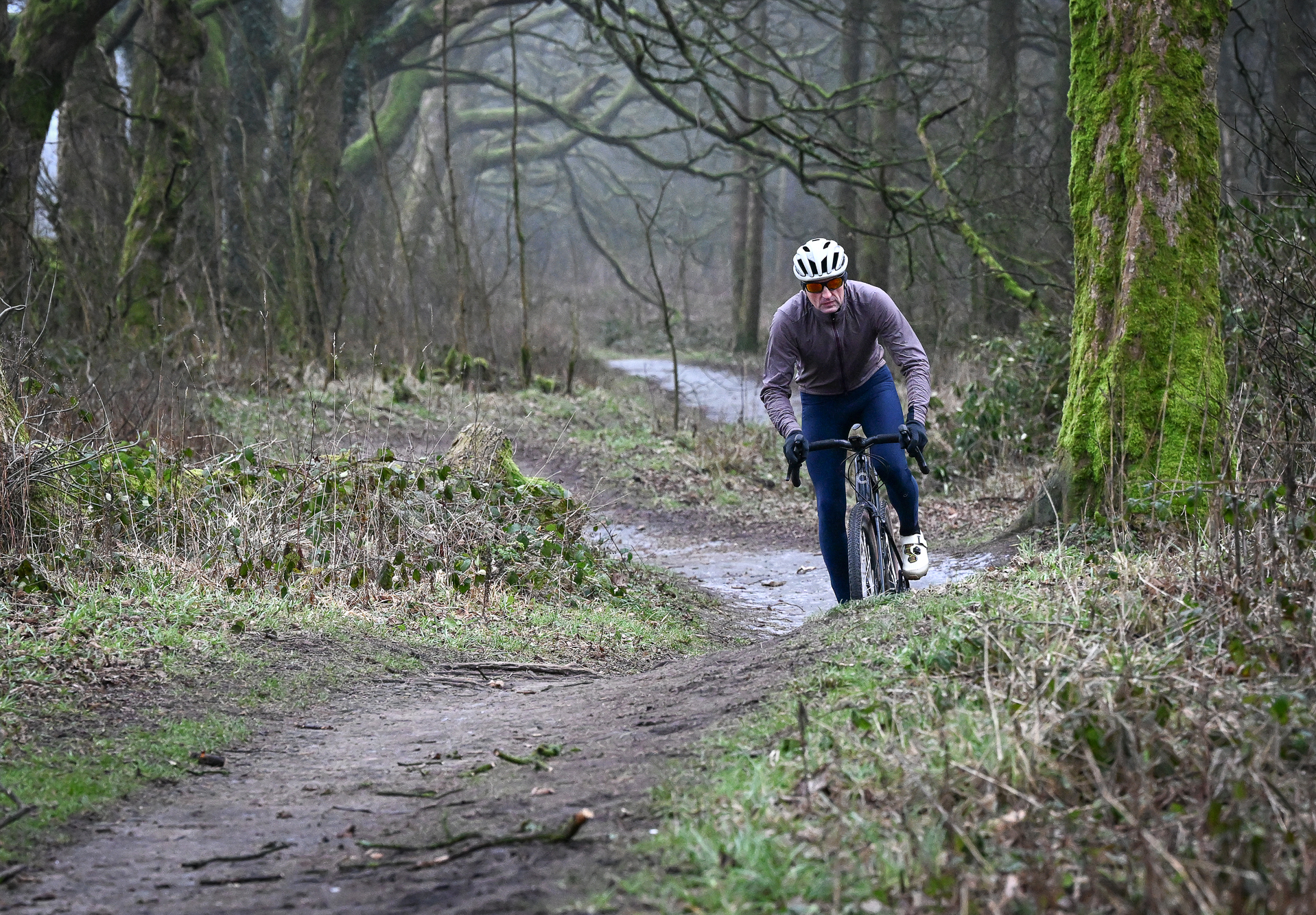 Man wearing a purple jacket, navy tights and a pale helmet riding a black gravel bike towards the camera on a muddy track
