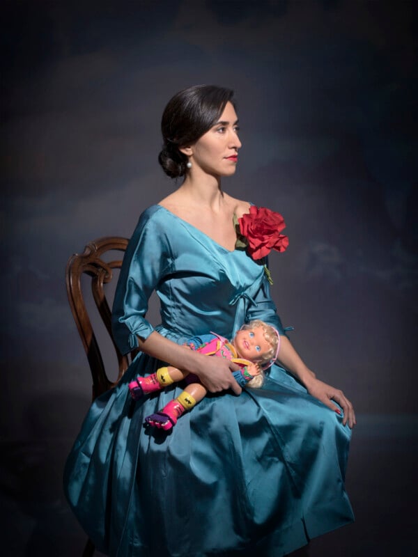 A woman in a teal satin dress with a large red flower sits on a wooden chair, holding a colorful toy doll in her lap, against a dark, softly lit background.