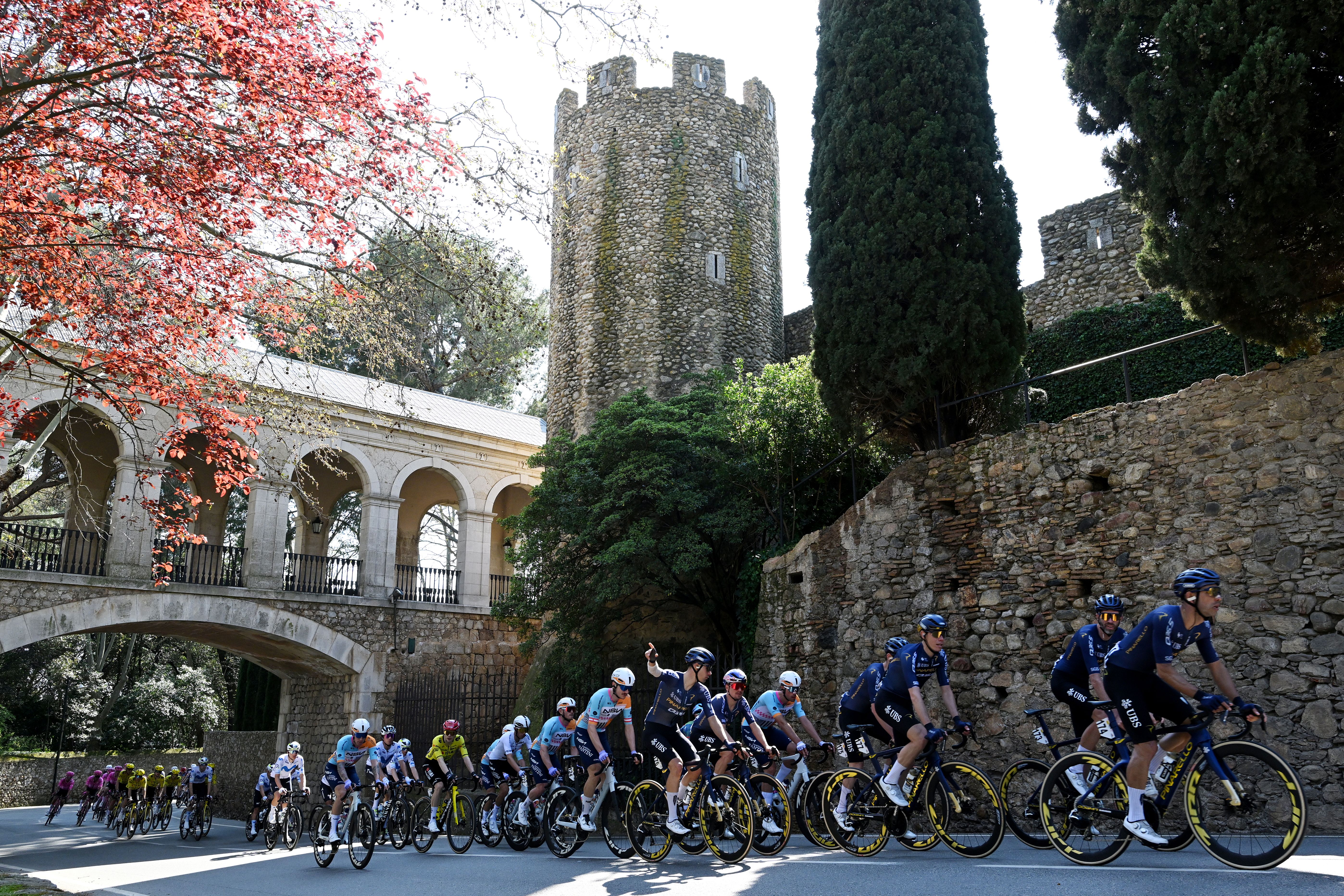 The peloton riding from left to right under an ancient bridge and passed a blossom tree on stage 2 of Volta a Catalunya 2026