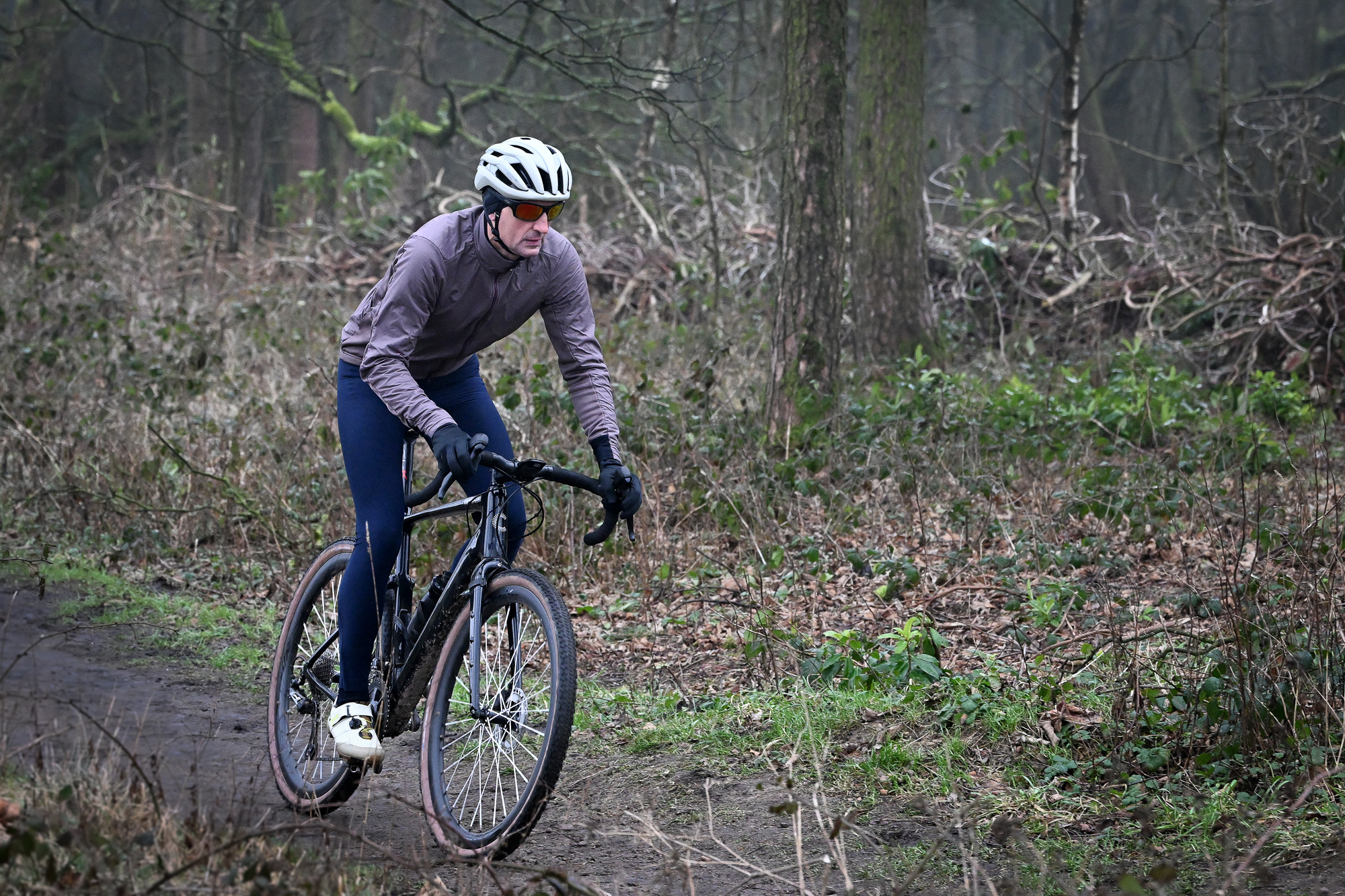 Man wearing a purple jacket, navy tights and a pale helmet riding a black gravel bike 3/4 towards the camera on a muddy track